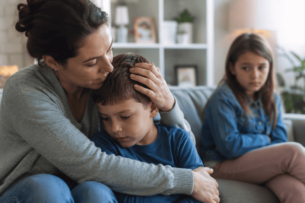 Mother comforting a young child on a couch while an older sibling sits in the background looking sad and left out.